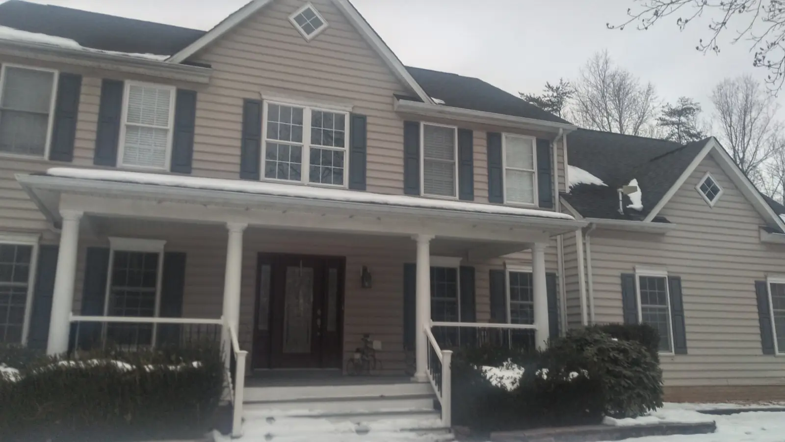 Two-story home with beige siding, dark shutters and a white-railed front porch in light snow.