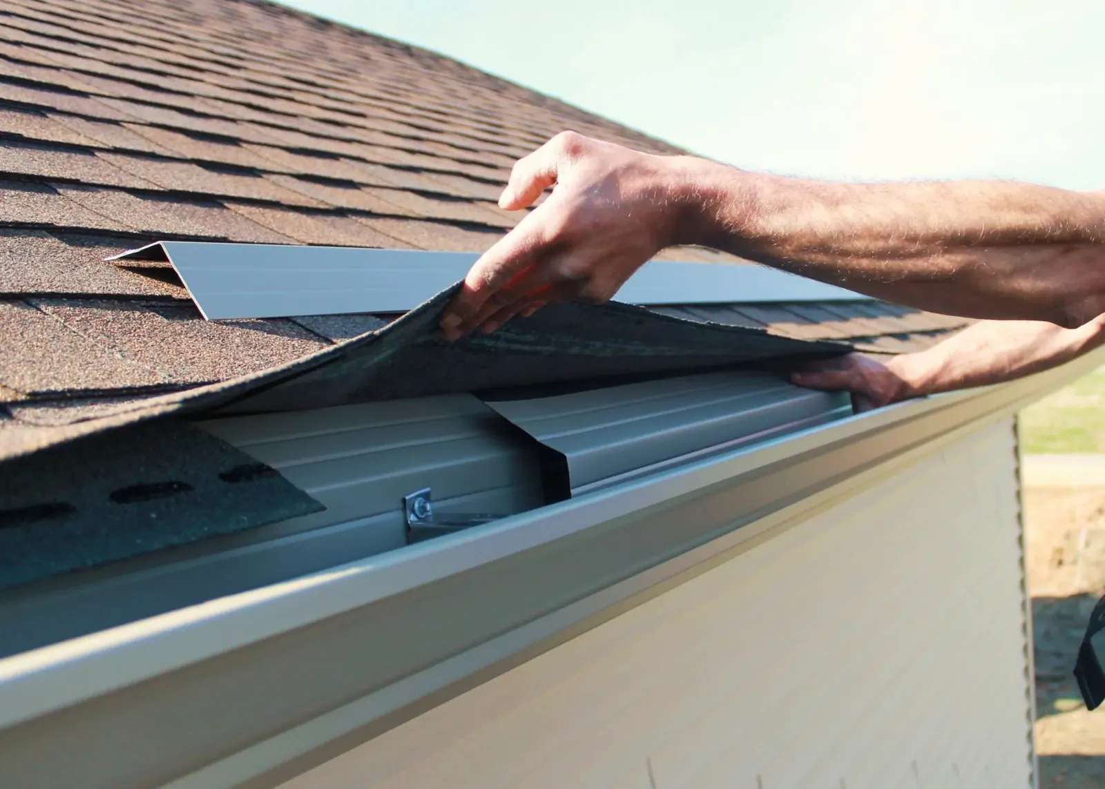 Hands fitting a metal gutter guard under the edge of a shingle roof above a white gutter.