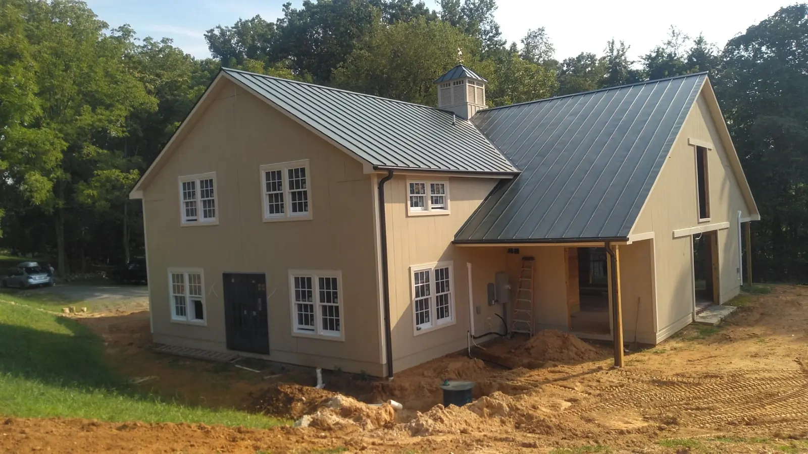Barn-style home with tan siding and a dark green standing seam roof, still under construction.