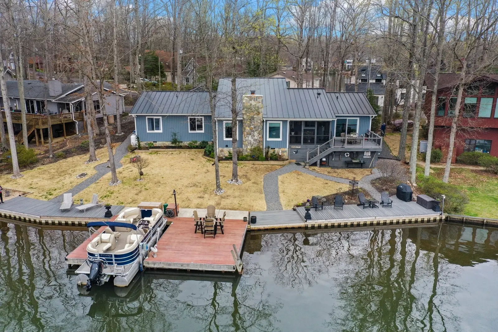 Blue-gray lake house with a dark metal roof, stone chimney, dock and pontoon boat.
