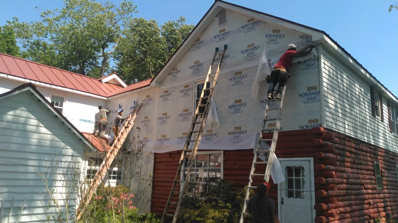 Crew on ladders installing house wrap on a two-story exterior beside a red metal roof.
