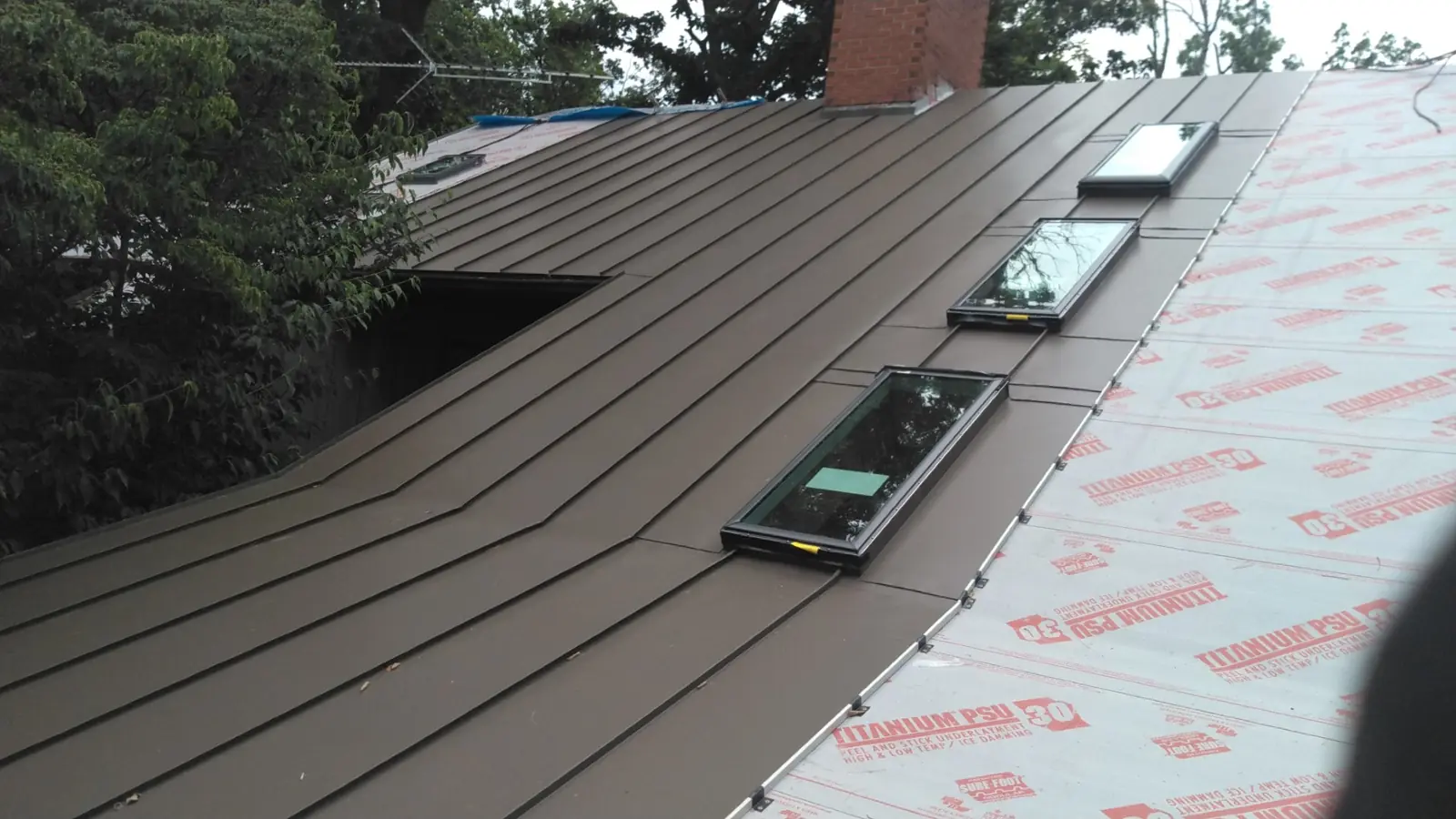 Brown standing seam panels around three skylights over red-printed underlayment.